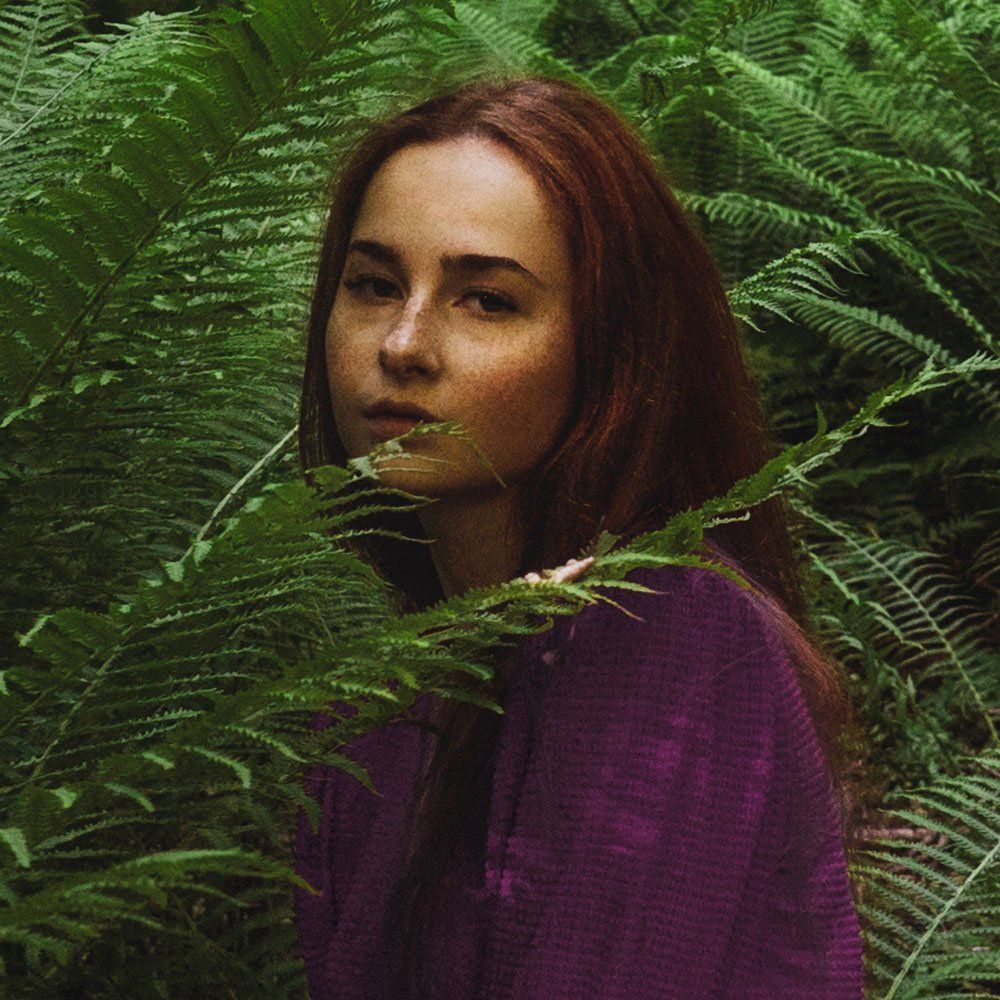 Woman in a purple sweater standing among lush green ferns, looking at the camera.