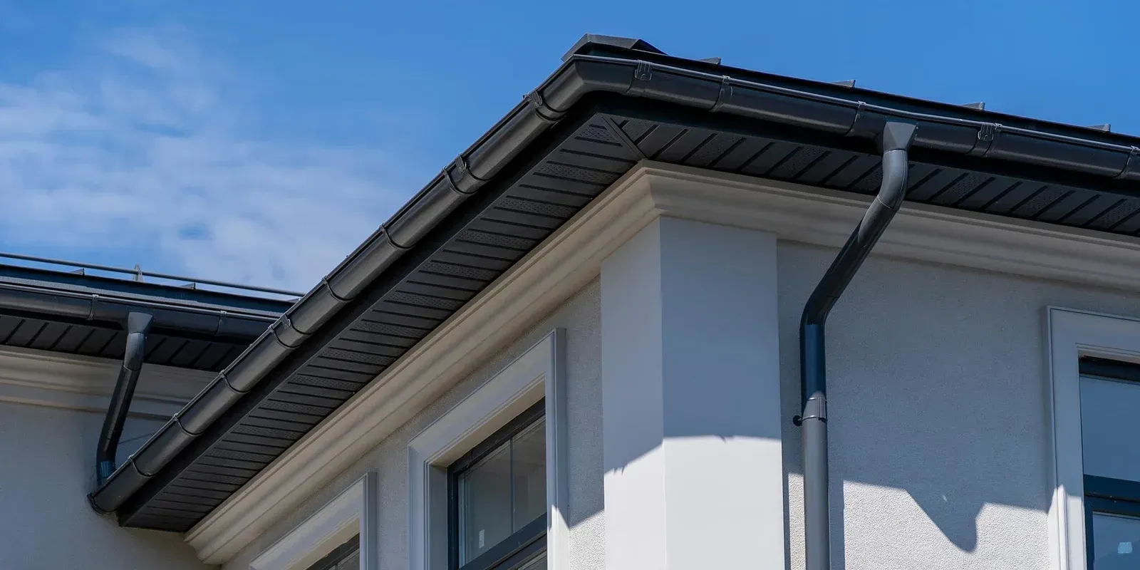The roof of a white house with a black gutter and a blue sky in the background.