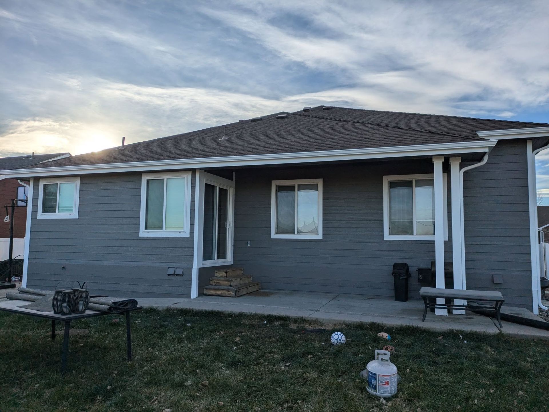 Gray single-story house with covered porch at sunset, with a grill and bench in the yard.