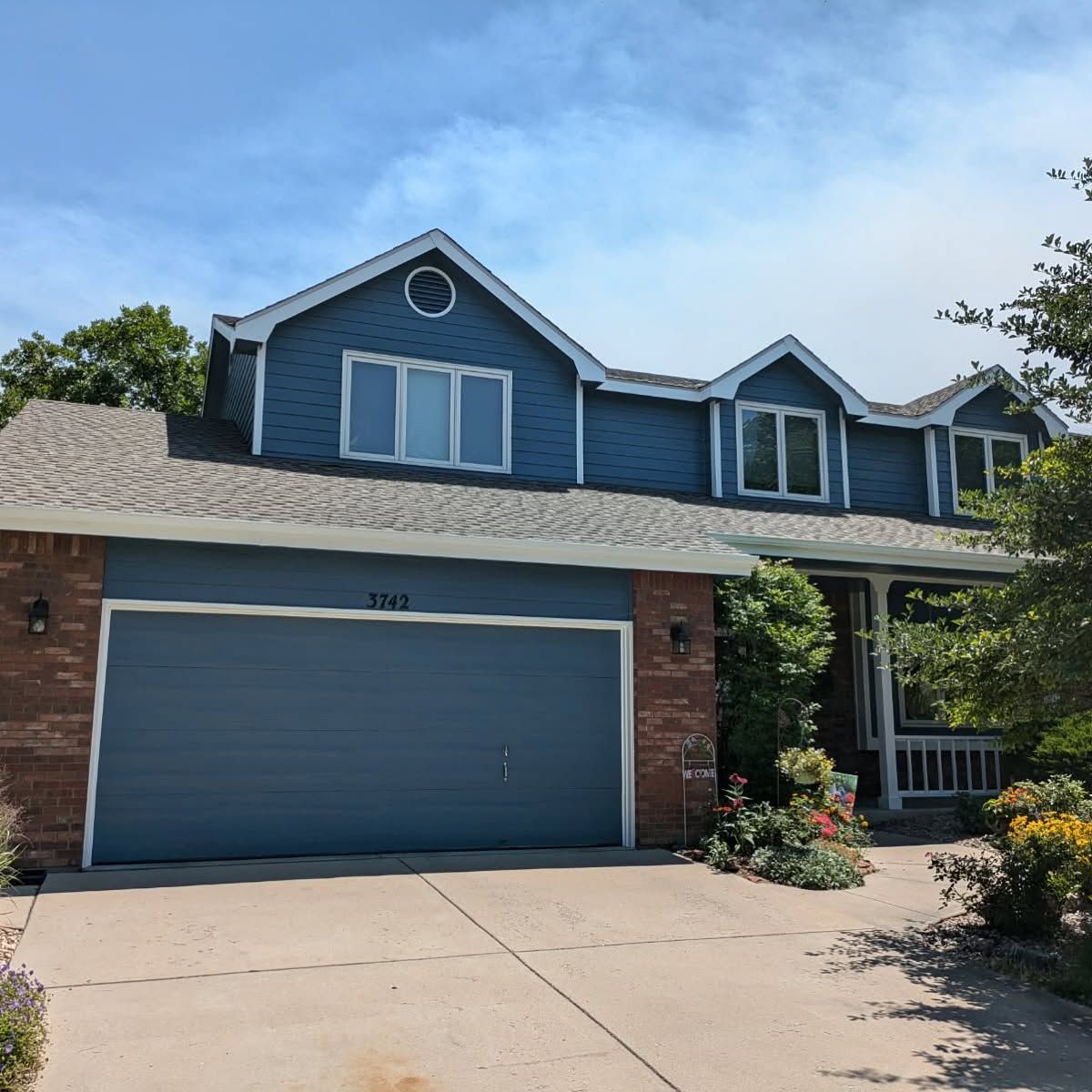 Blue two-story suburban house with brick garage, driveway, and front porch under a sunny sky