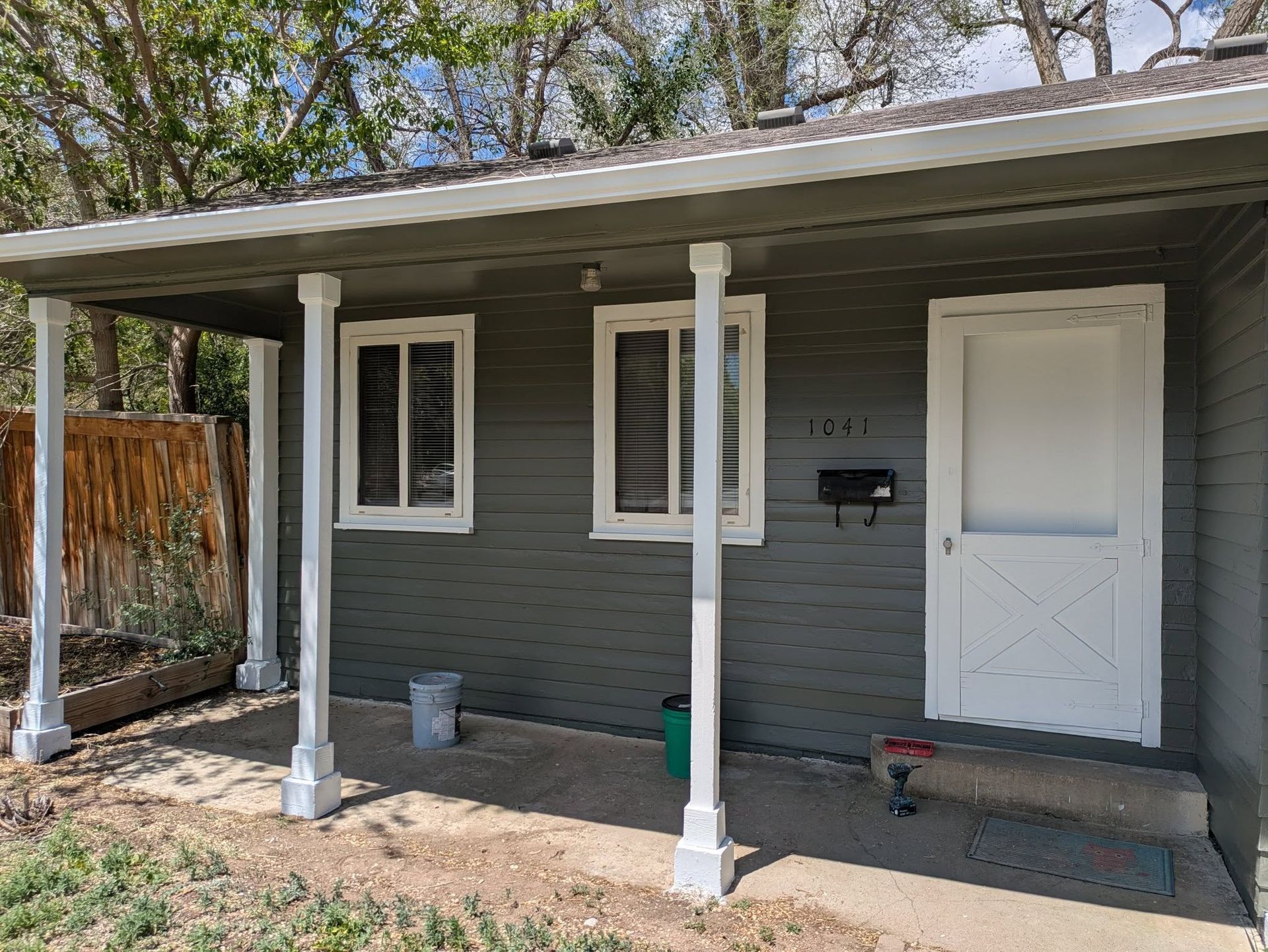 Small gray house porch with white columns, front door, windows, and a mailbox in daylight