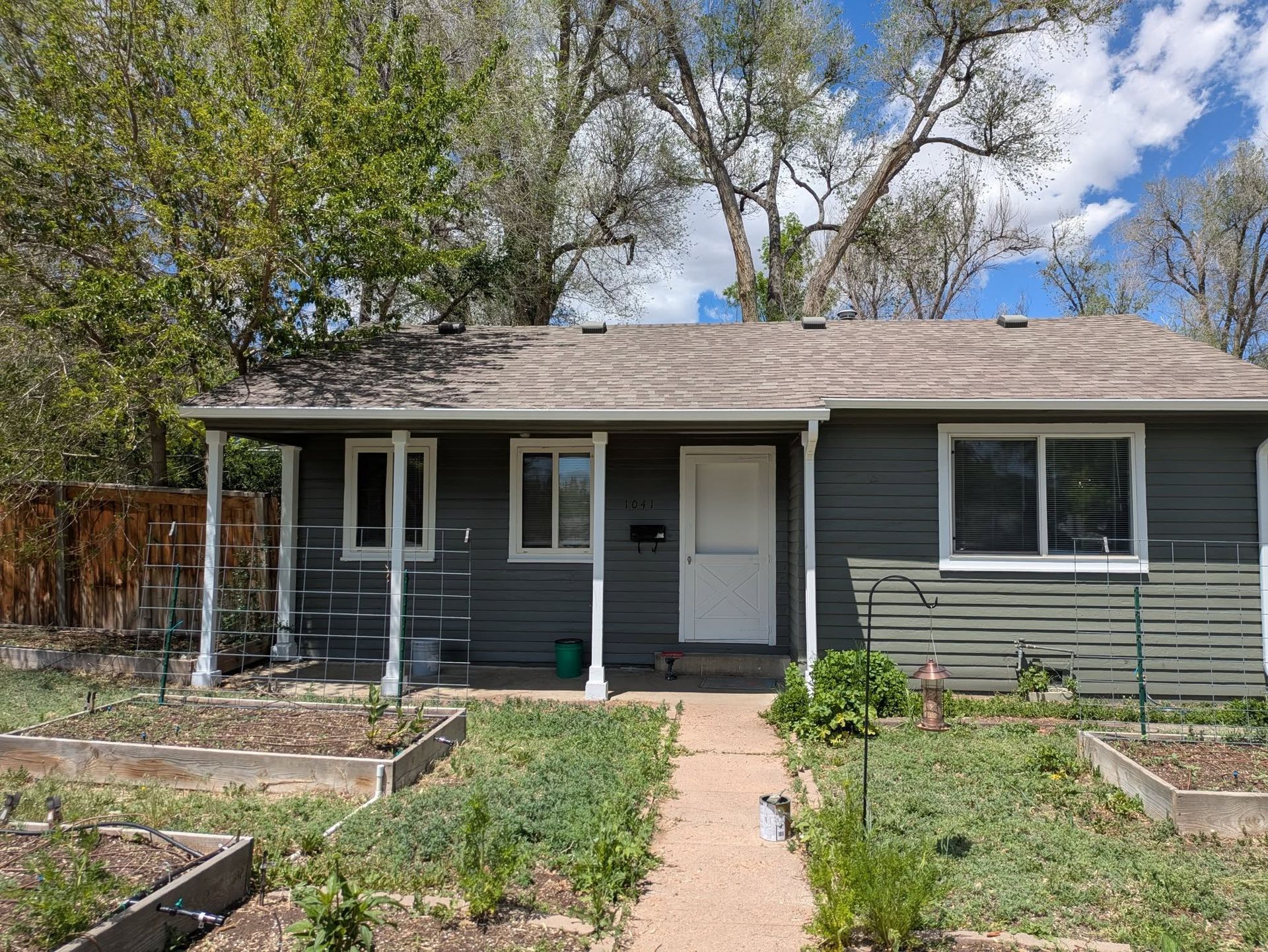 Small gray house with a front porch, white door, and walkway, surrounded by a garden under a blue sky.
