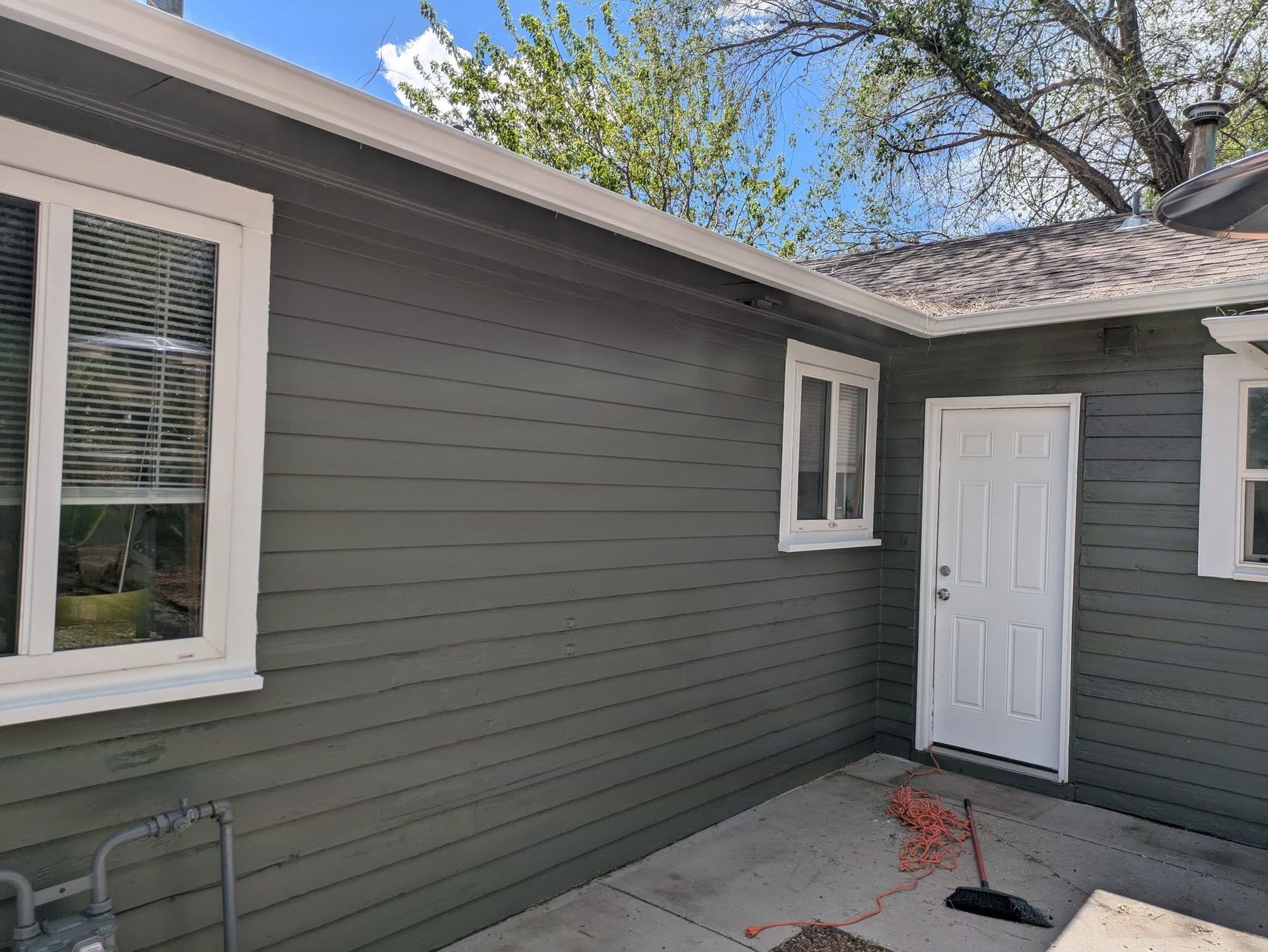 Gray house exterior with white-trim windows, a side door, and a small concrete patio under trees