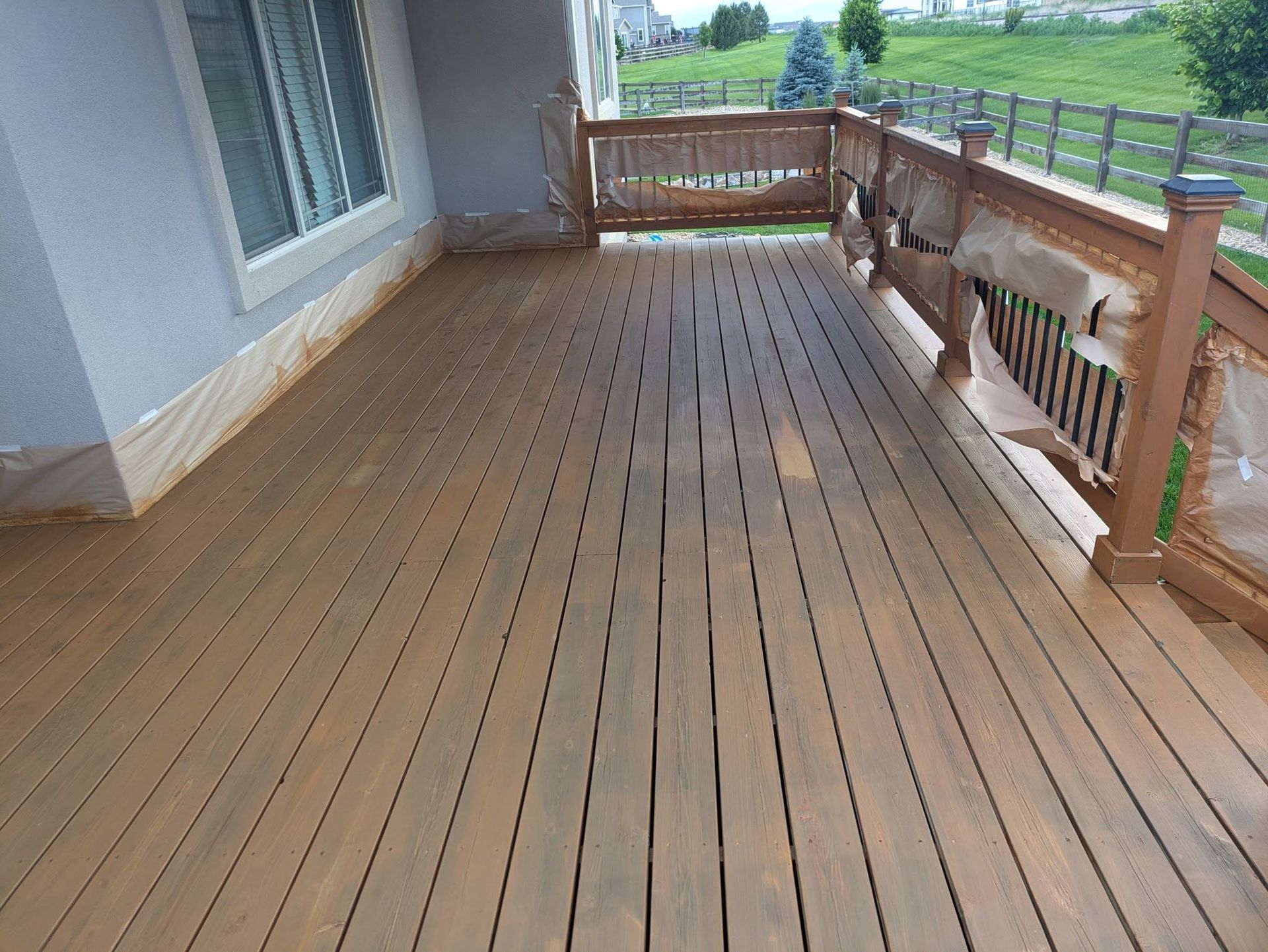 Wooden outdoor deck with railing beside a house, overlooking a green yard and field.