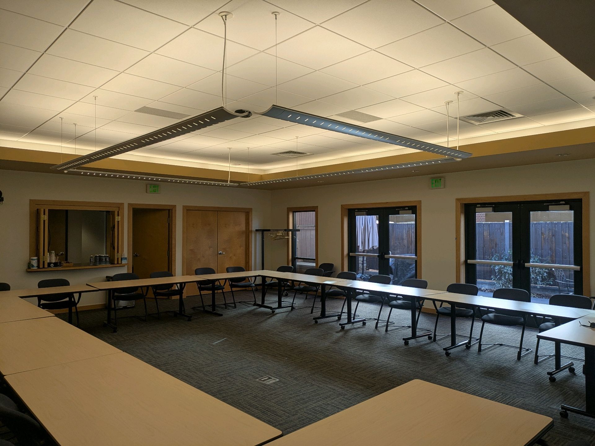 Empty conference room with U-shaped tables, chairs, and ceiling lights around a carpeted floor.