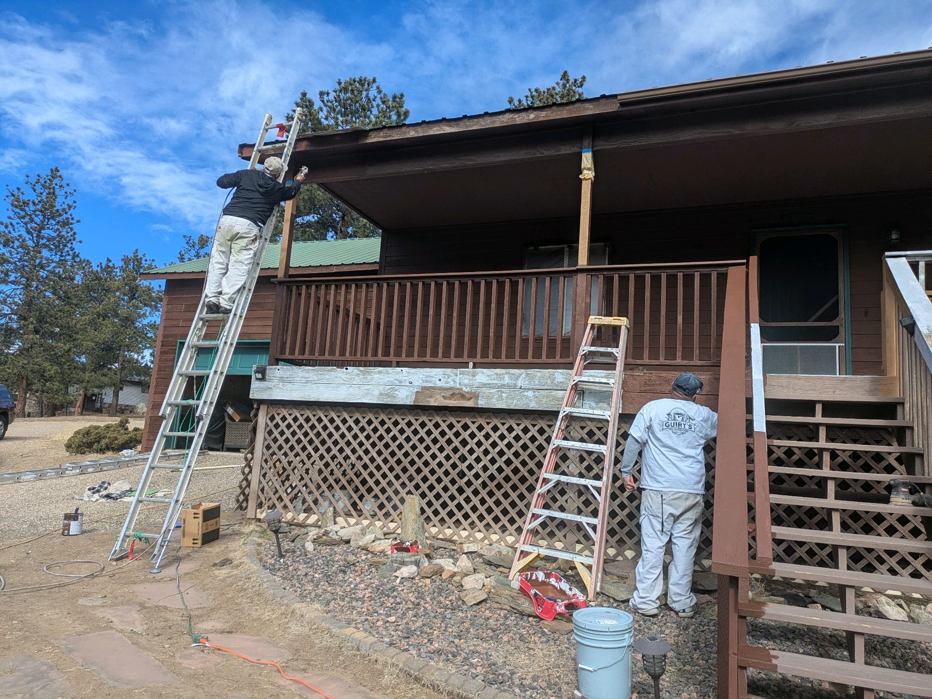 Two workers on ladders repairing a cabin porch and roof exterior.