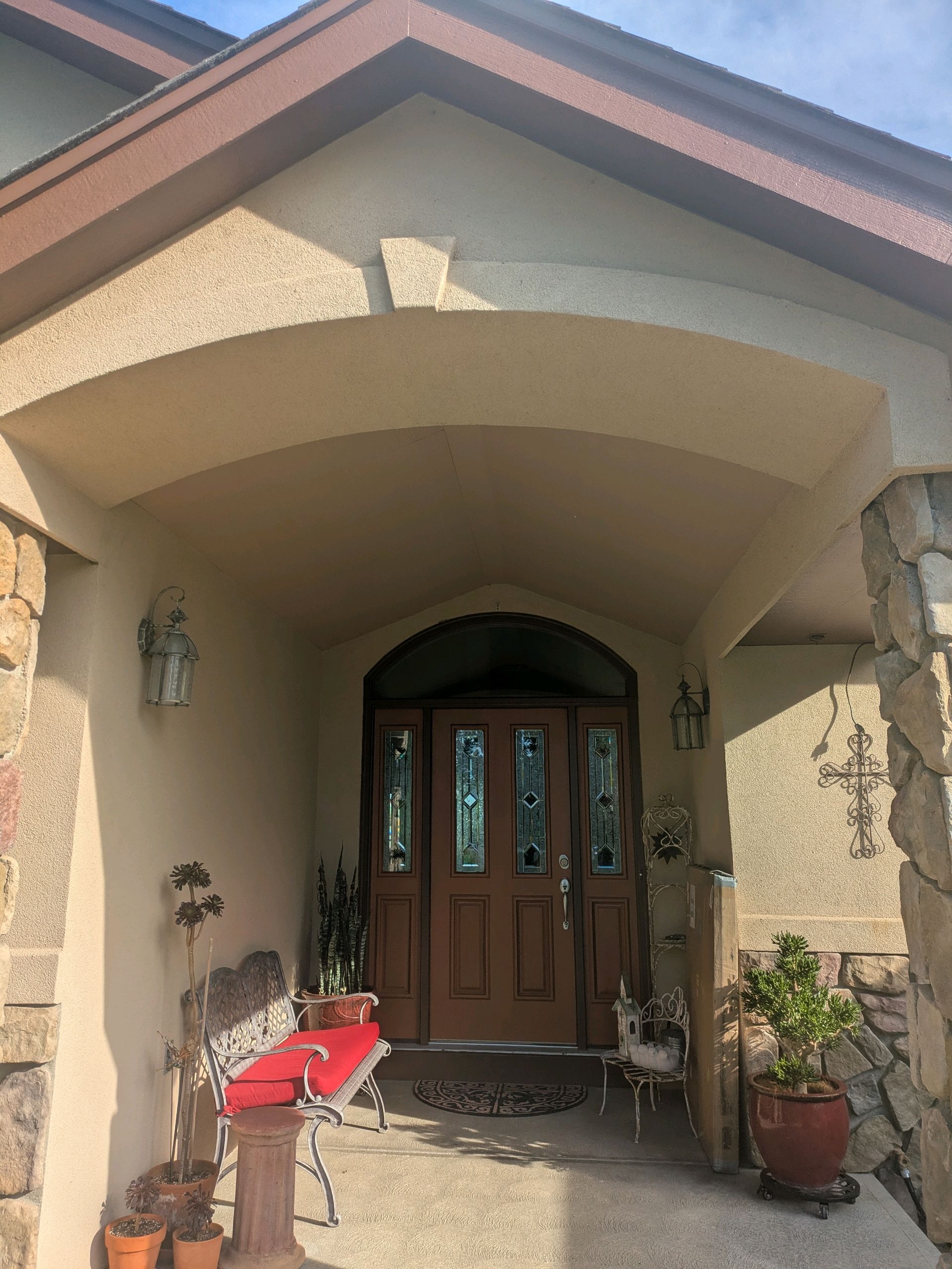 Front entrance of a stucco house with arched porch, wooden door, wall lights, and potted plants on the steps