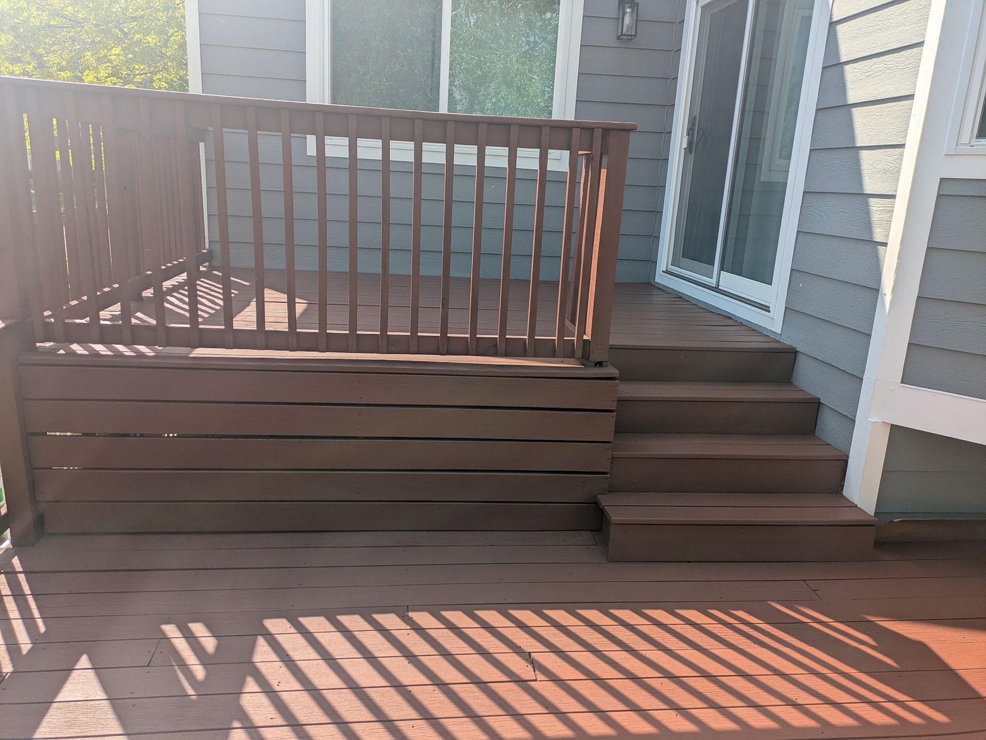 Wooden deck steps leading to a raised patio with brown railing beside a house.
