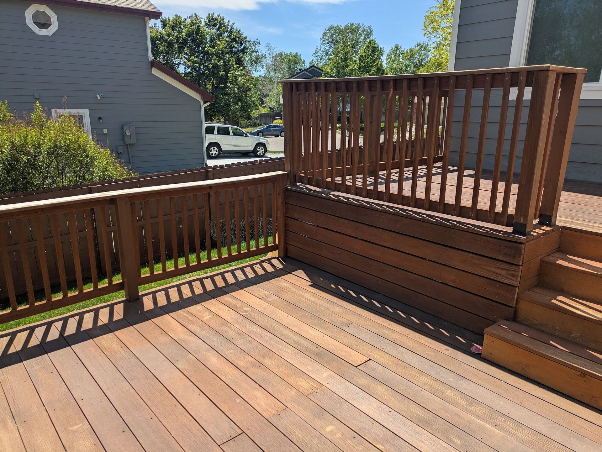 Wooden deck with railing and steps beside a house, overlooking a sunny yard and street.
