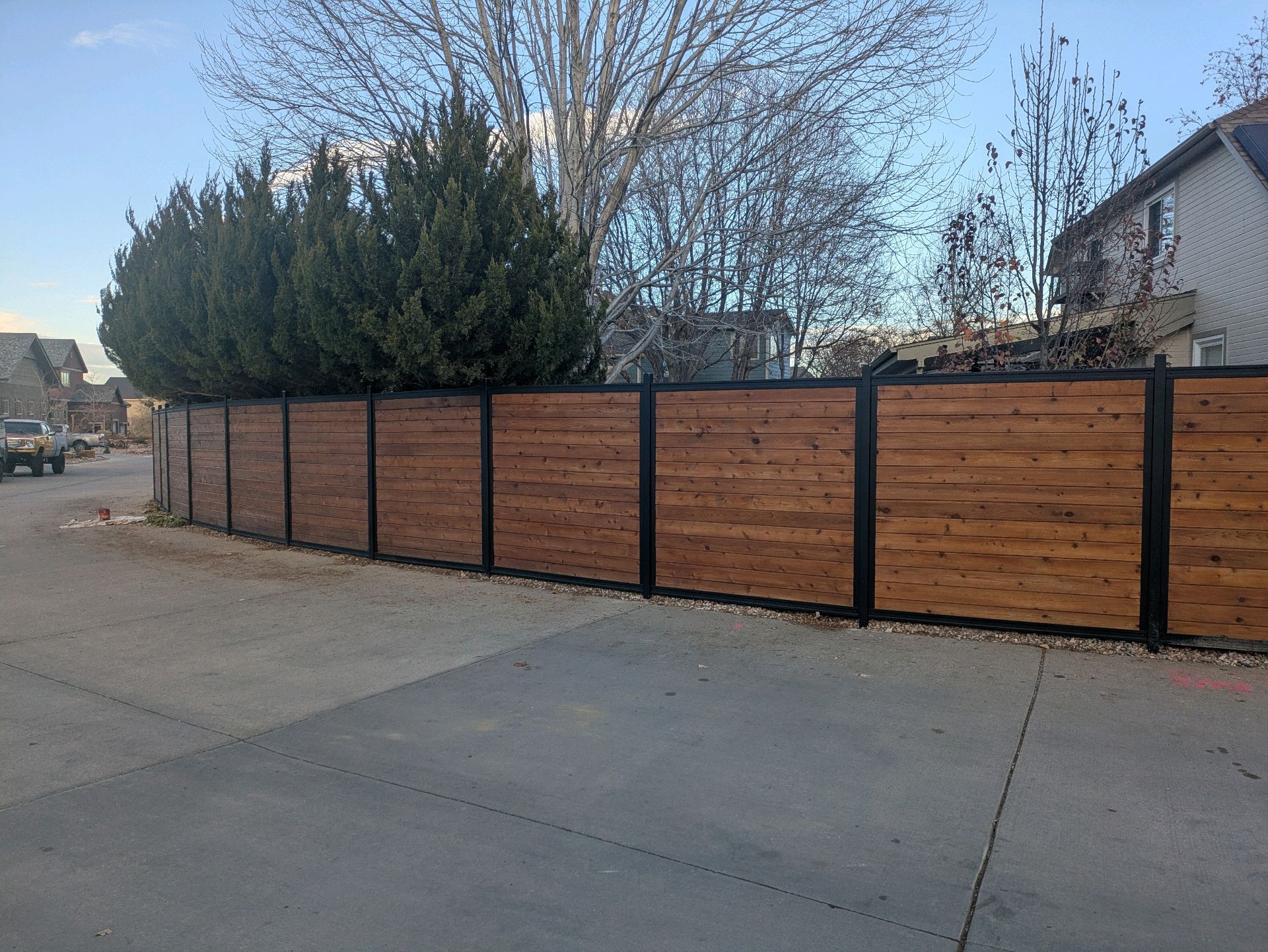 Wooden fence with black posts lining a quiet roadside beside leafless trees and a house