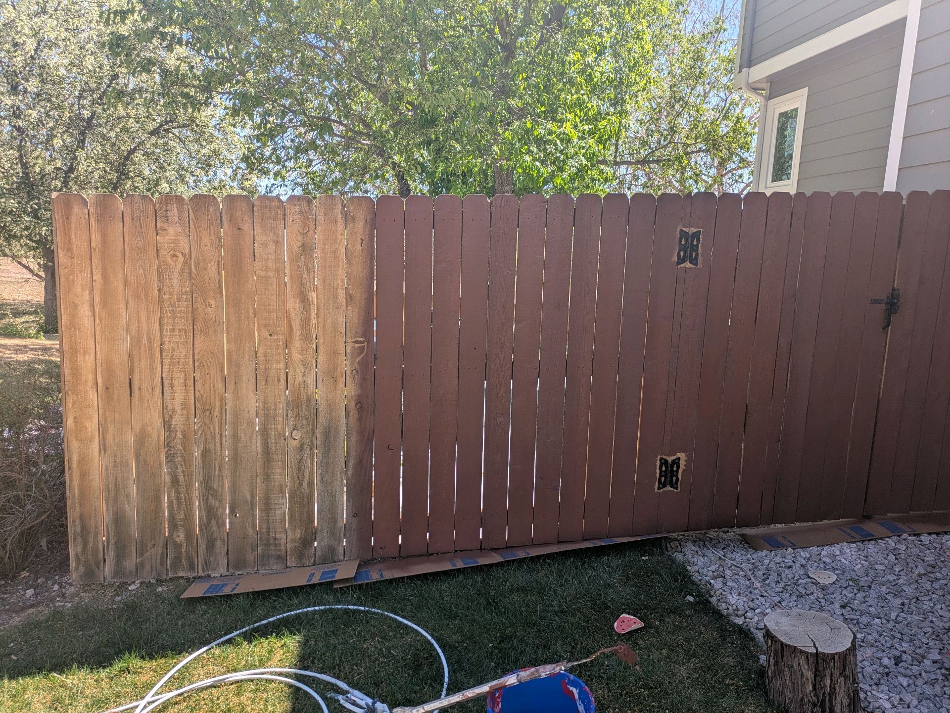 Wooden backyard fence with one weathered panel, grass, and garden hose in front, beside a house and trees