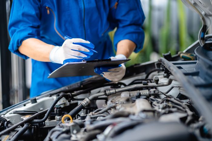 Mechanic in blue uniform inspecting a car engine with a clipboard in a garage