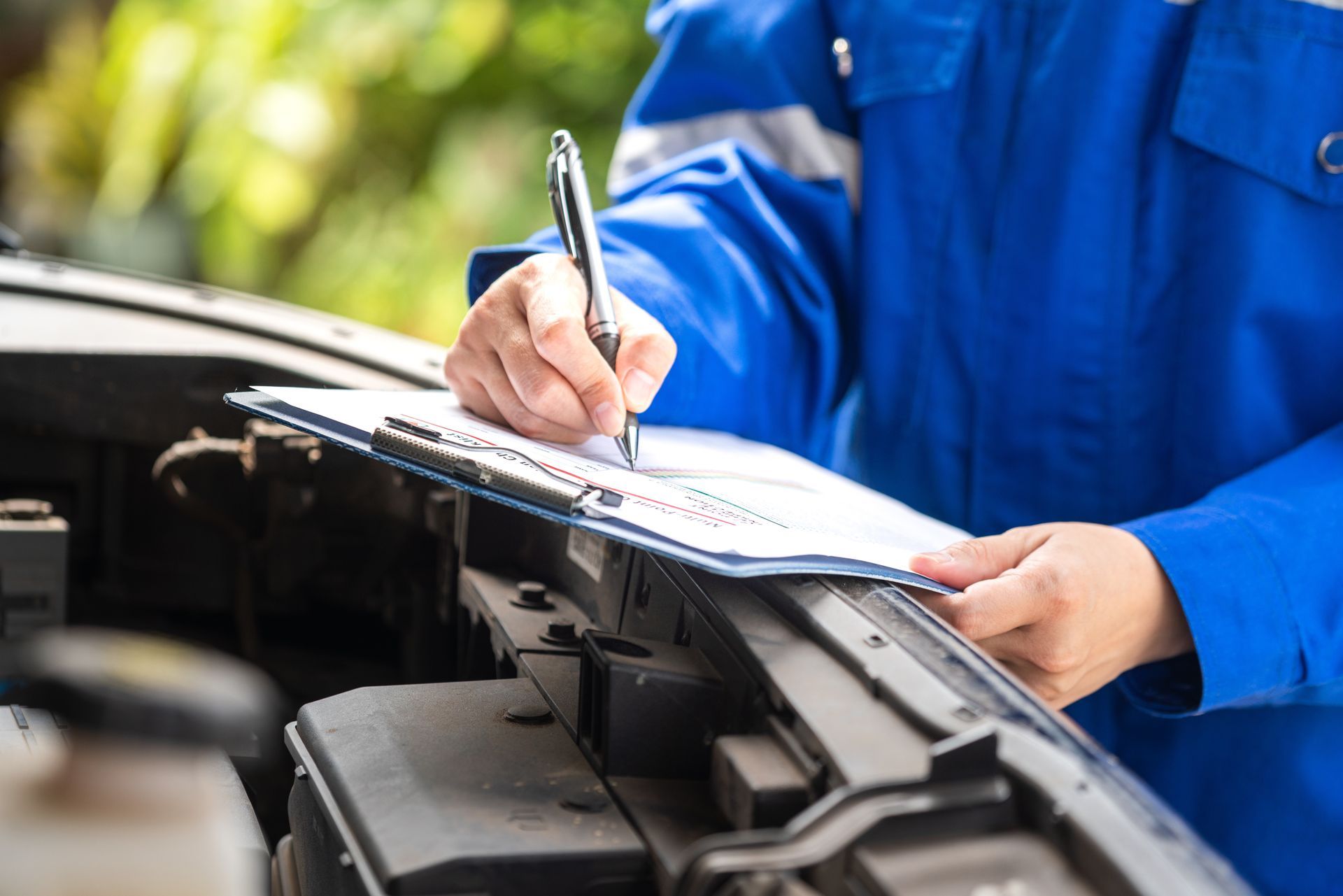 Mechanic in blue uniform writing on a clipboard beside an open car hood