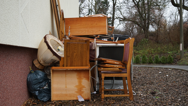 Stack of wooden furniture and boxes piled beside a wall outdoors, with a bag and chair on the ground.