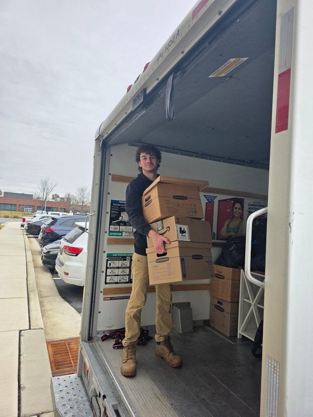Person carrying stacked boxes in the back of a delivery truck at a parking lot.