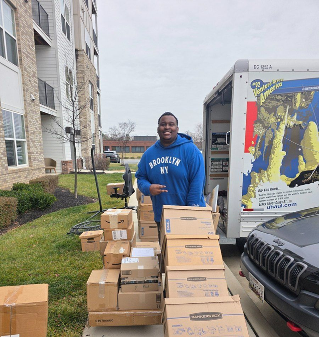 Man standing outside apartment building beside stacked cardboard boxes and moving truck, wearing blue sweatshirt.