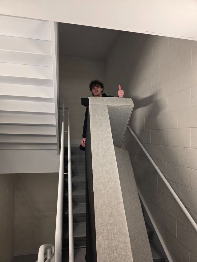 Person on indoor staircase beside a tall gray structure, giving a thumbs-up.