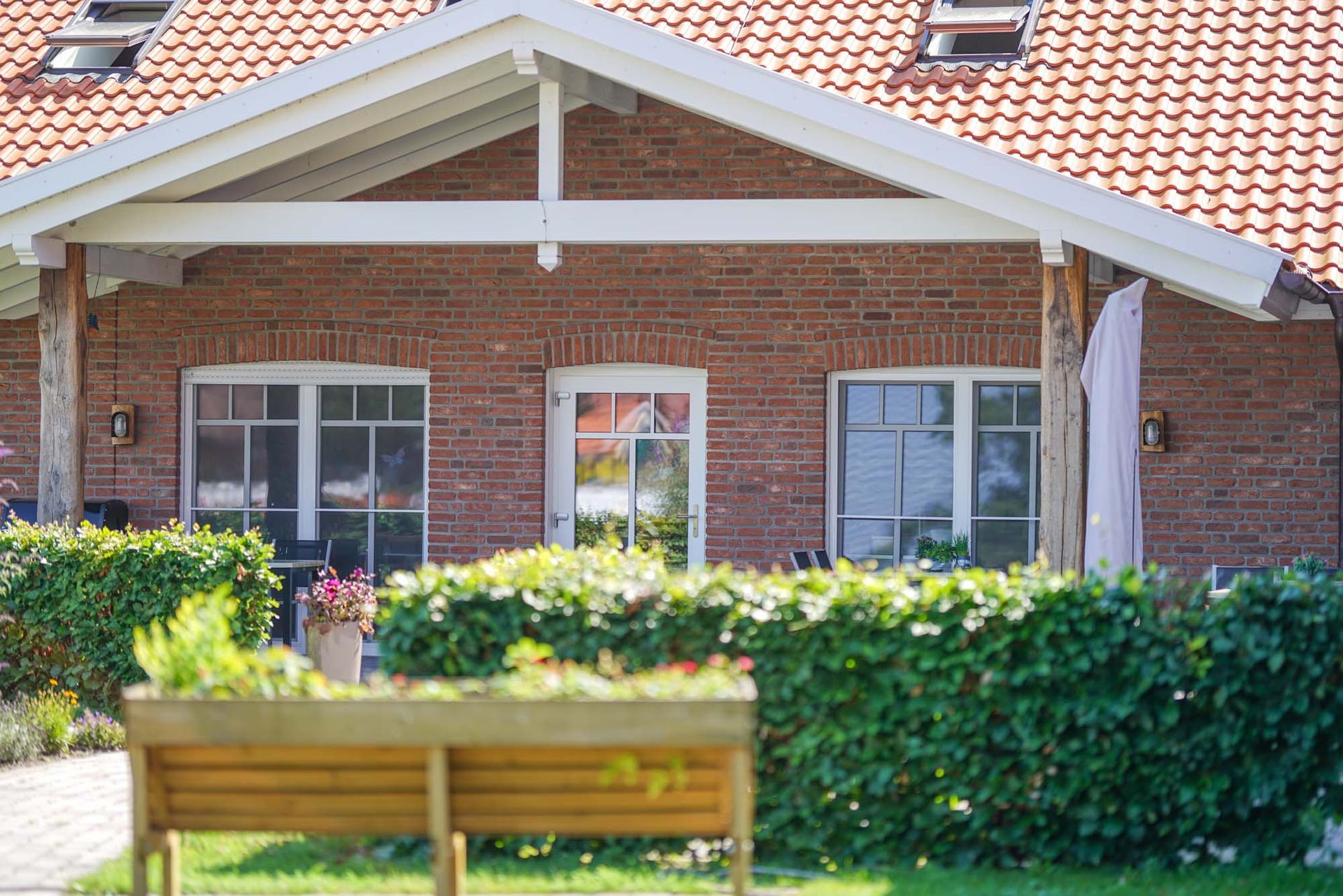 Ein Backsteinhaus mit weißem Dach und einer Holzbank davor. Tagespflege Up Deele vom Garten aus