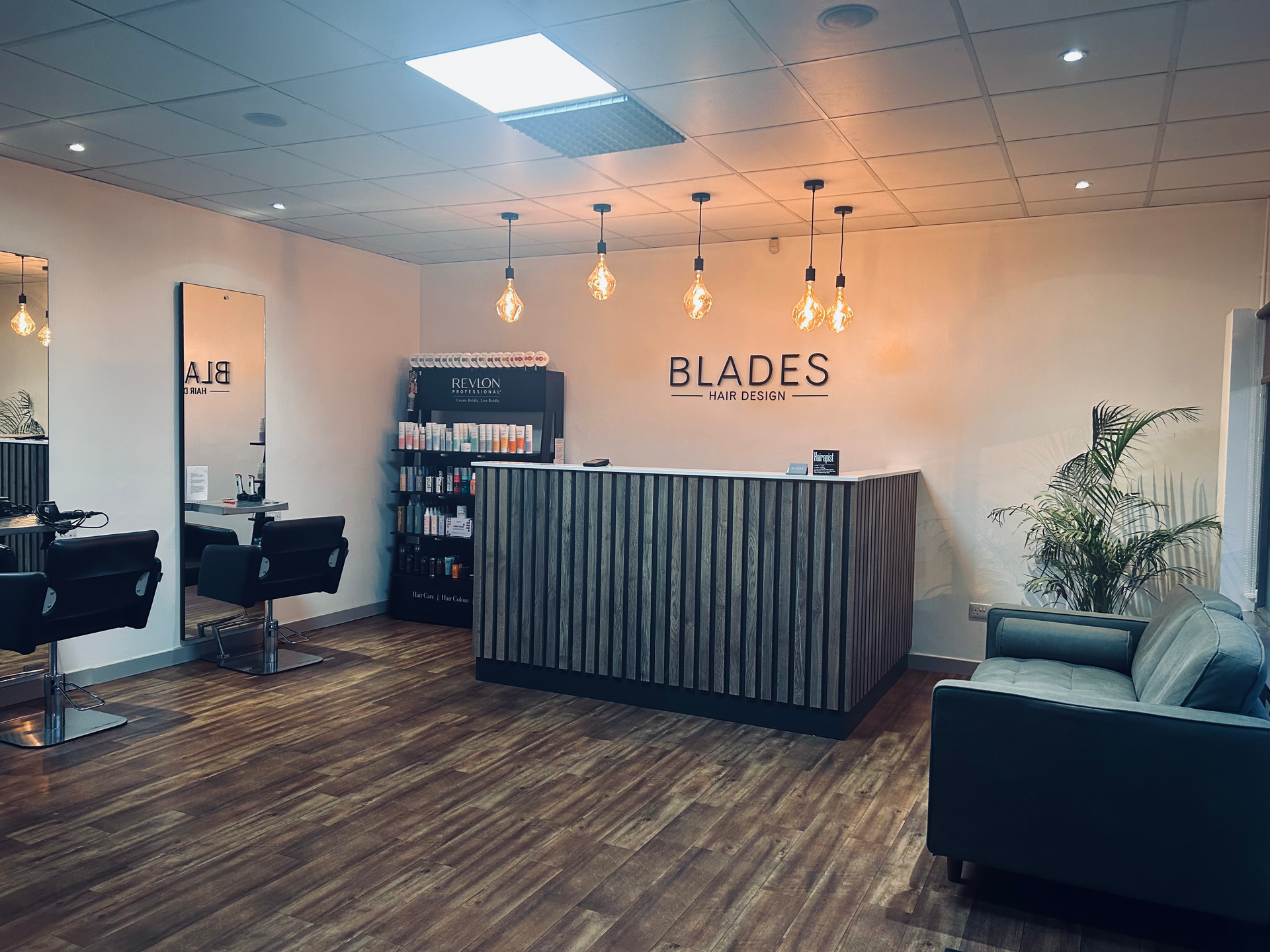 Modern salon reception with striped desk, black sofa, pendant lights, and wooden floors under a “BLADES” sign