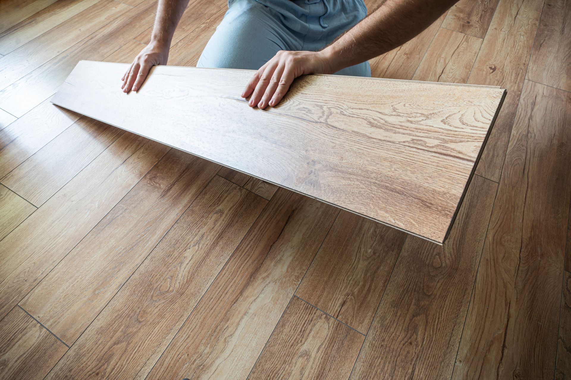 A person kneeling on a floor, placing a light-colored wood plank into a laminate flooring installation. A person kneeling on a floor, placing a light-colored wood plank into a laminate flooring installation.