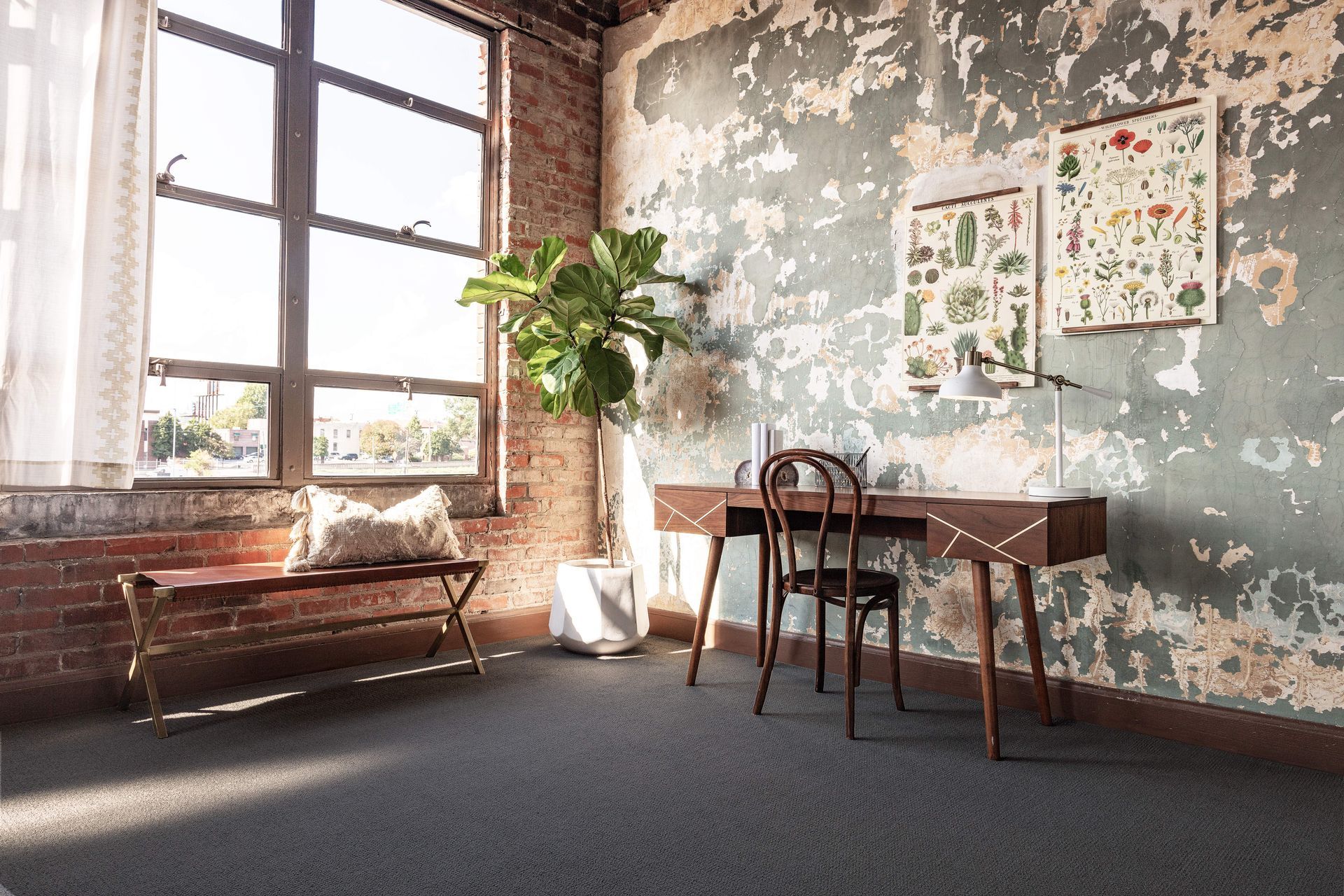 Sunlit loft with large windows, rustic desk, chair, plant, and worn brick-and-plaster walls