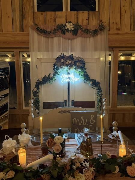 Wedding reception sweetheart table with floral arch, candles, and monogrammed backdrop in a rustic hall