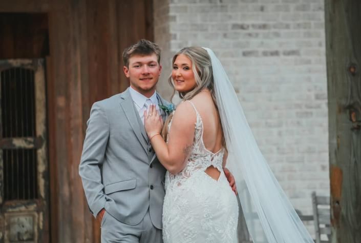 Bride and groom posing together outdoors, with the bride in a white lace dress and veil, and the groom in a light gray suit