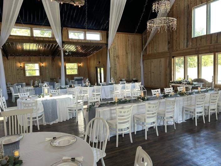 Elegant rustic banquet hall with long white-draped tables, white chairs, chandeliers, and large windows