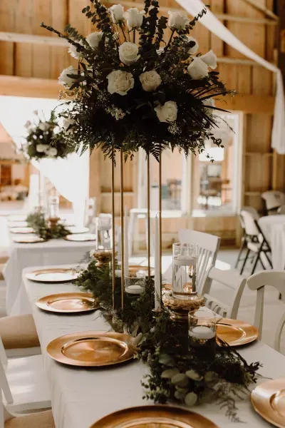 Elegant wedding reception table with gold plates, white linens, and a tall floral centerpiece in a barn venue