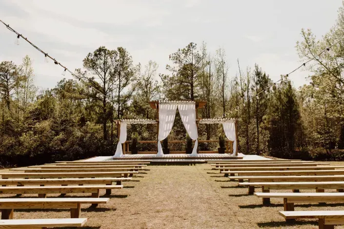 Outdoor wedding altar with white drapes, benches, and string lights in a wooded clearing