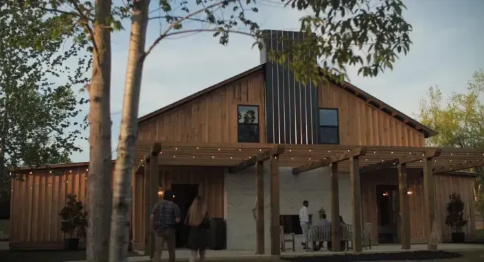 Wooden barn-style house with a covered porch and people standing outside under trees at dusk