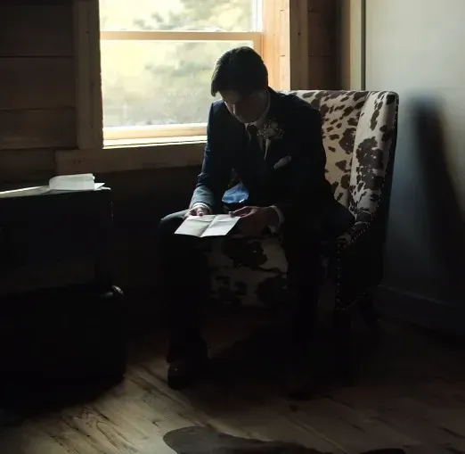 Person sits in a chair by a window, reading a book in a dim room.