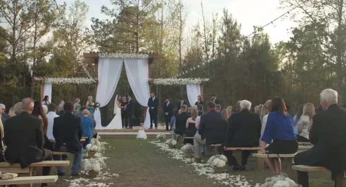 Outdoor wedding ceremony with guests seated on benches along a flower-lined aisle under trees