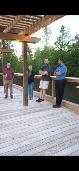 Four people stand on a wooden deck under a pergola, surrounded by trees at sunset.