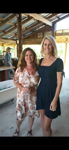 Two women standing and smiling indoors under a rustic wooden roof