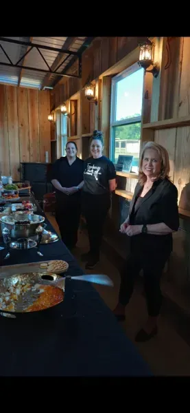 Three people standing beside a buffet table in a rustic wood-paneled room with tall windows and hanging lights