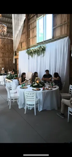 Guests seated at a round table in a rustic barn-style venue with white drapery and hanging greenery