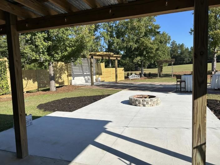 Covered outdoor patio with concrete seating area, stone fire pit, and trees in a sunny park setting