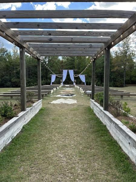 Garden walkway under a wooden pergola leading to white chairs and draped fabric in the distance