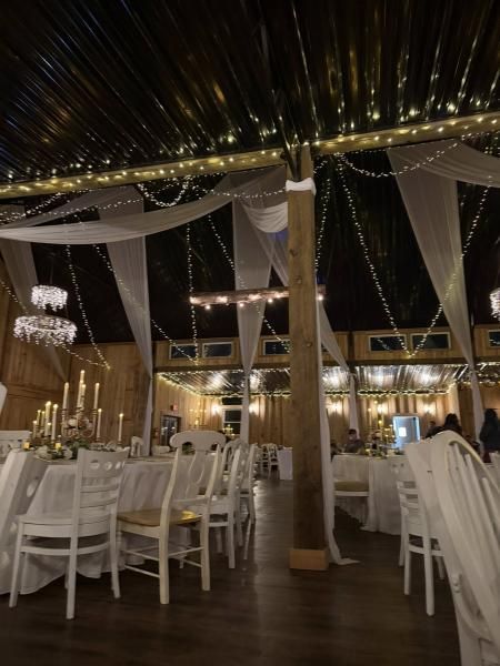Elegant outdoor wedding reception with white chairs, draped fabric, string lights, and wooden tables under a dark canopy