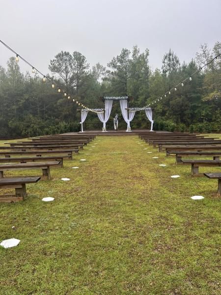 Outdoor wedding ceremony setup with wooden benches, white draped arch, and string lights on a grassy lawn.