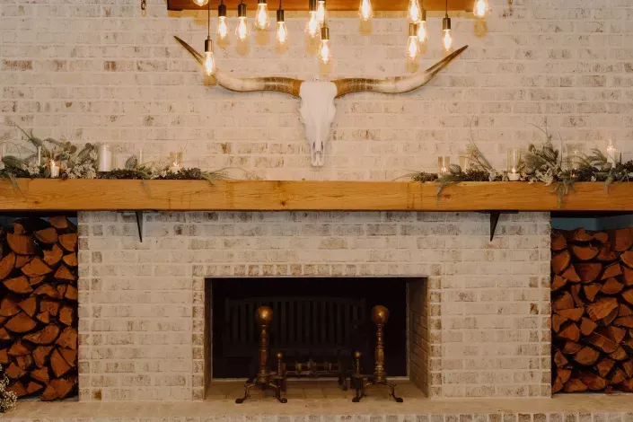 Rustic fireplace with white brick surround, stacked firewood, and a deer skull chandelier above