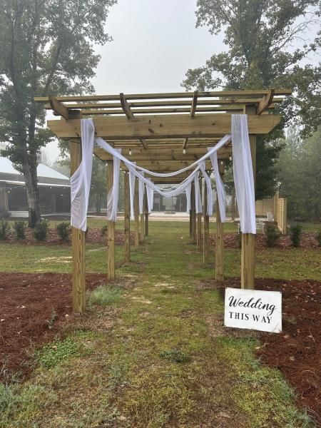 Outdoor wedding aisle under a wooden arch with white drapes and a “wedding this way” sign.