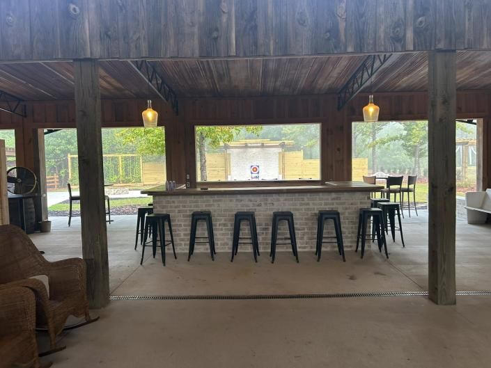 Open-air pavilion with a long bar, black stools, and hanging lights overlooking a wooded view