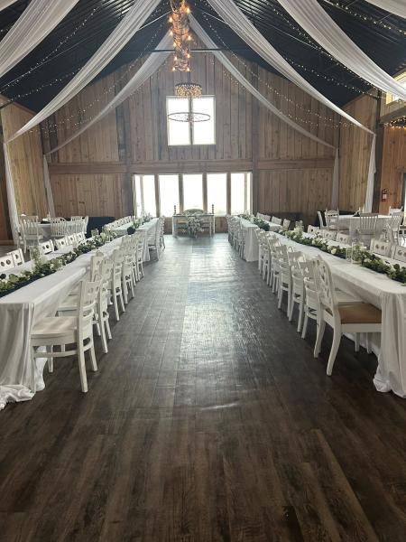 Barn venue set for a wedding reception, with long white tables, chairs, and draped fabric overhead.