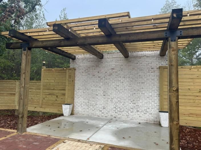 Wooden pergola over a concrete patio with white brick wall and fenced backyard panels