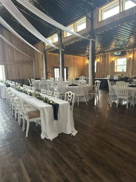 Empty rustic banquet hall with long white-draped tables, white chairs, and dark wood floors under draped ceiling decor