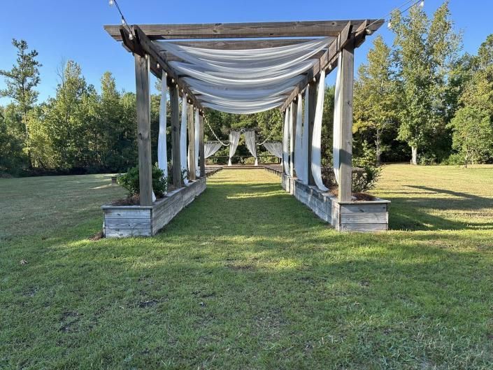 Wooden pergola draped with white fabric, centered on a grassy lawn with trees in the background