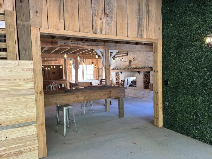 Rustic open interior with wooden beams, a counter, stools, and a green hedge wall on the right