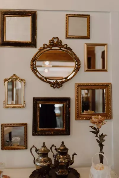 Decorative wall of assorted gold-framed mirrors and art above a small table with silver teapots and a vase.